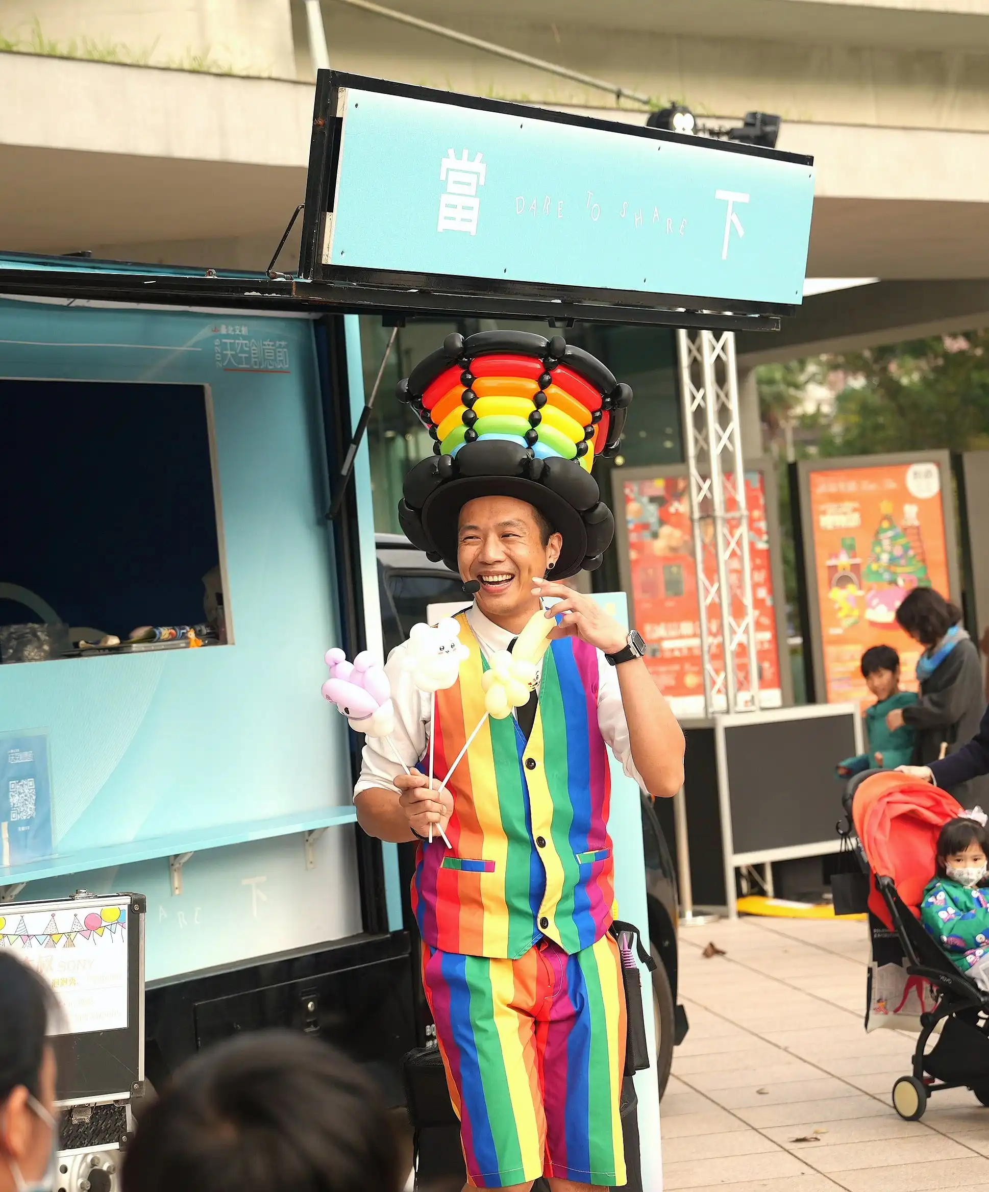 氣球大叔手拿造型氣球搭配彩虹帽，在臺北文創活動現場笑容滿面
