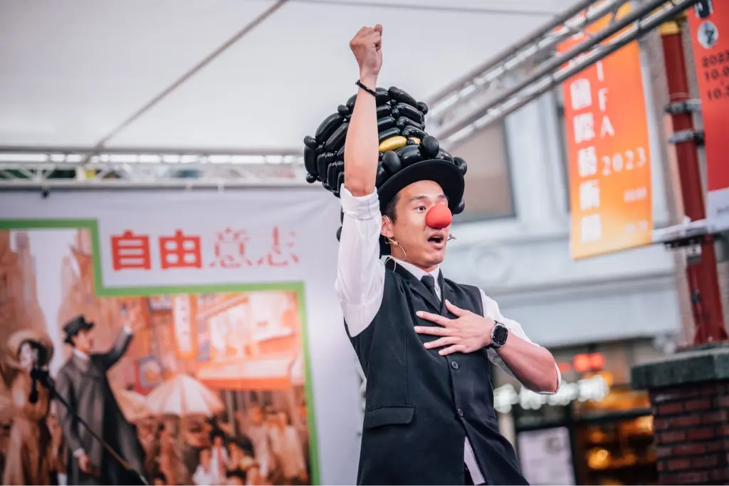 大稻埕國際藝術節街頭表演,氣球表演者戴著黑氣球帽與紅鼻,在自由意志主題布幕前誇張演出