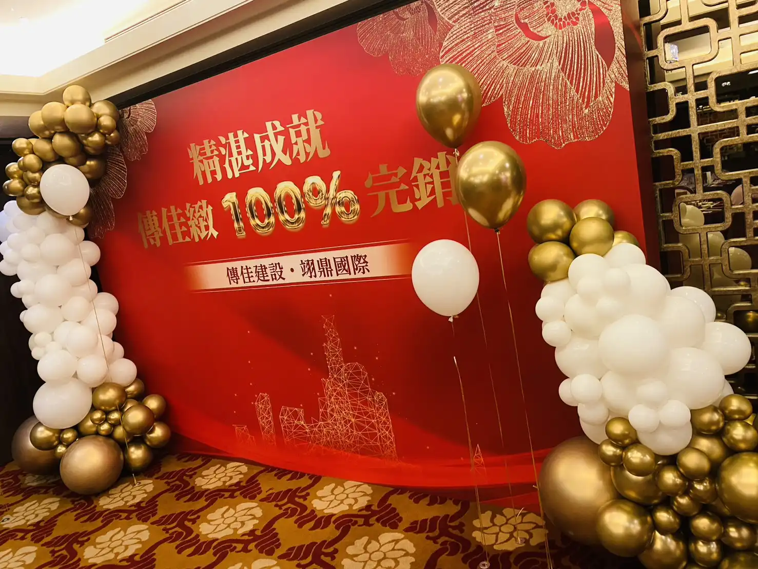 傳佳建設完銷慶功宴金白色氣球佈置細節，紅底背板上印有「精湛成就 傳佳緻100%完銷」字樣