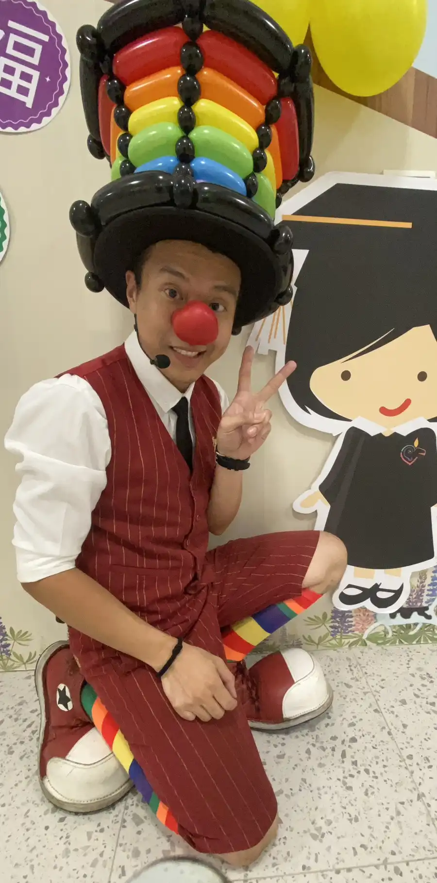 氣球大叔Sony戴著彩虹氣球帽和紅鼻子,在桃園中堅幼兒園畢業典禮後台與卡通立牌合影,比出勝利手勢