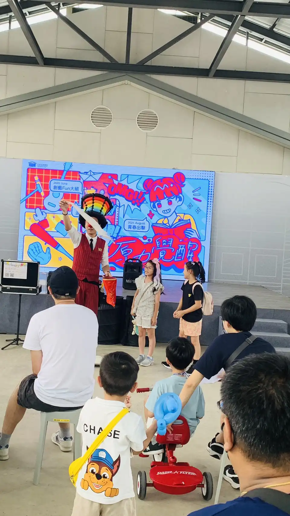 氣球大叔戴彩虹帽於桃園中原文創園區舞台上與兩位小朋友互動，背景為活動主視覺